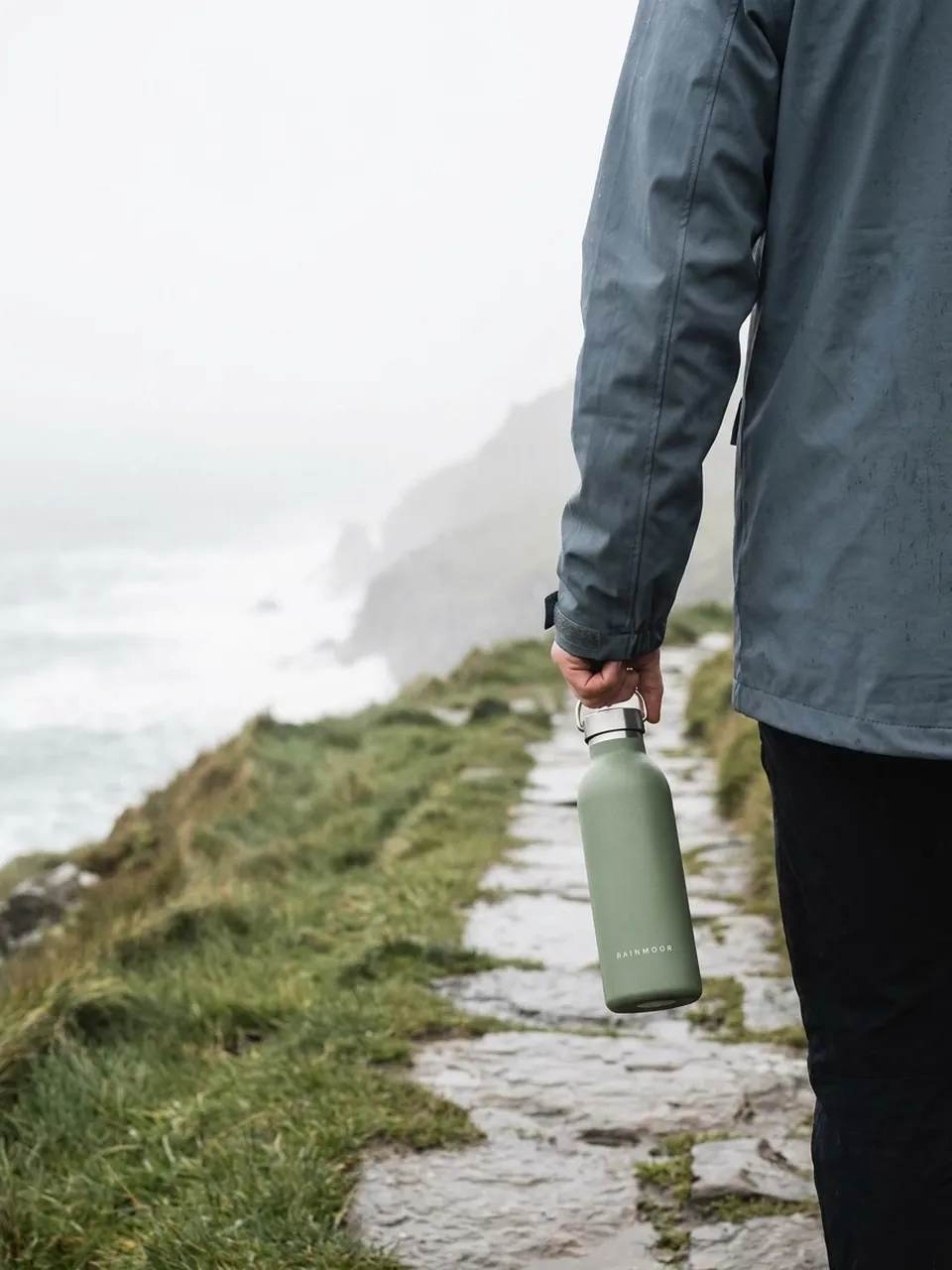 AI-generated example lifestyle-angle ad for Rainmoor, a fictional drinkware brand: a hiker holds the sage-green bottle on a misty coastal trail after rain