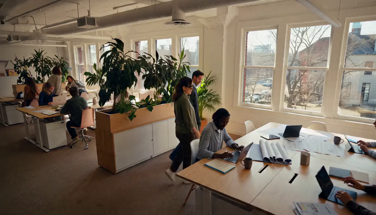 Colleagues working and talking around a large table in a bright open-plan office with tall windows and plants