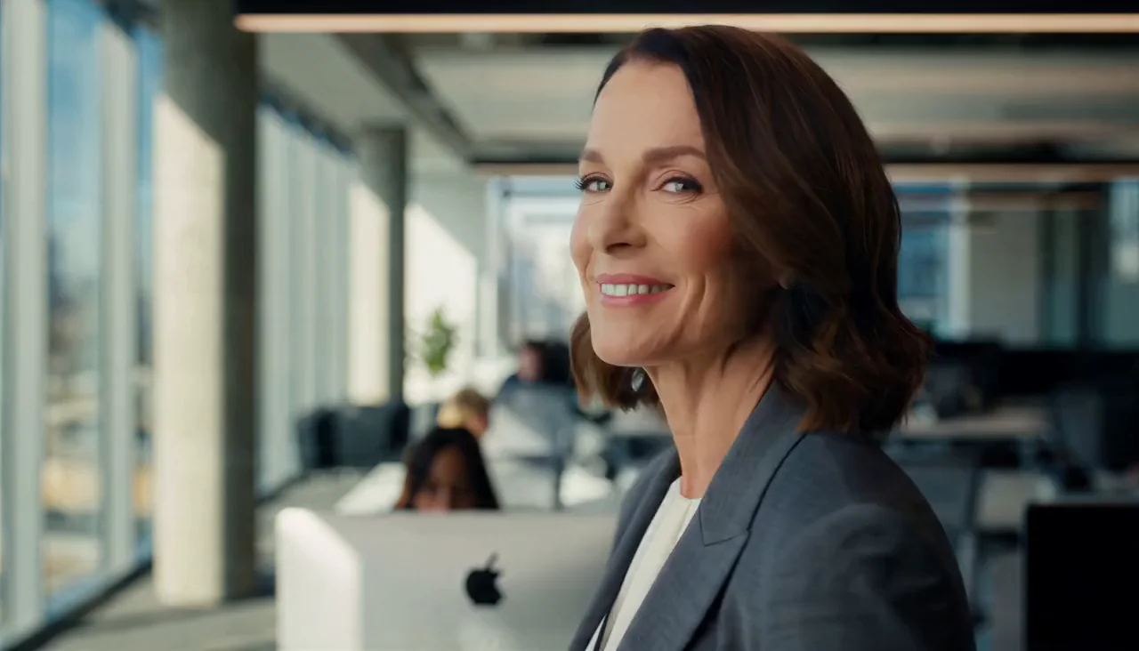 A leader in a grey blazer speaking directly to camera in a bright modern office, city visible through the windows behind her